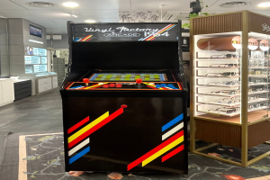Ein Geschäft mit einem zentralen Retro-Arcade-Automaten, einem Regal mit Gläsern auf der rechten Seite und Hintergrund-Elementen wie Schränken, einem Fernseher und anderen Gegenständen, mit Deckenlampen, die eine Retro-Atmosphäre schaffen.