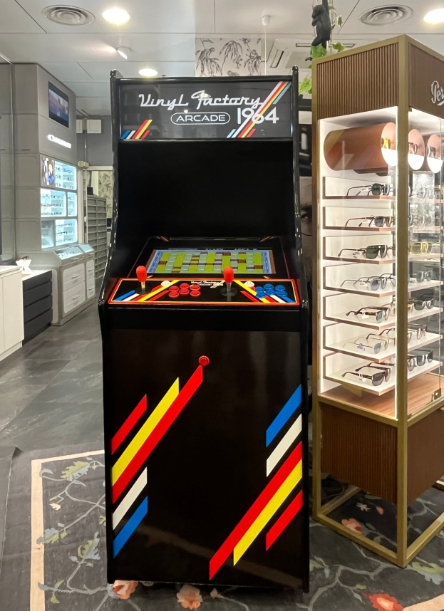 Ein Geschäft mit einem zentralen Retro-Arcade-Automaten, einem Regal mit Gläsern auf der rechten Seite und Hintergrund-Elementen wie Schränken, einem Fernseher und anderen Gegenständen, mit Deckenlampen, die eine Retro-Atmosphäre schaffen.