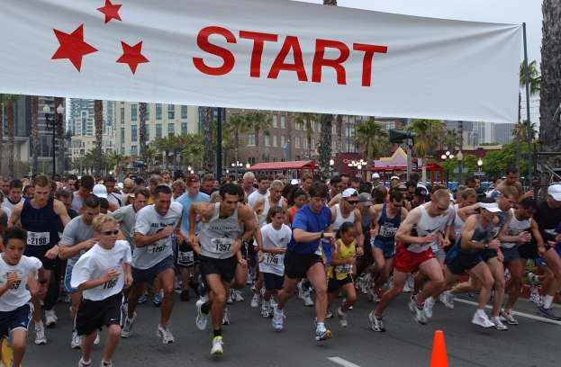 Gruppe von Läufern bei einem Marathon mit einem Verkehrskegel im Vordergrund und einer Fahne mit Text im Hintergrund, vor Bäumen, Laternenmasten, Gebäuden und einem klaren blauen Himmel.