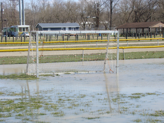 Ein Fußballtor steht in der Mitte eines überfluteten Feldes, umgeben von Gras, Wasser, Hütten, Pfosten, Bäumen, Fahrzeugen und einem klaren blauen Himmel.