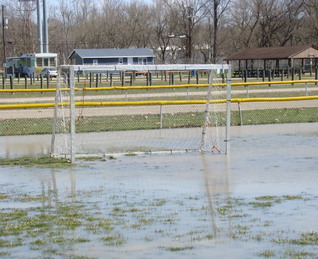 Ein Fußballtor steht in der Mitte eines überfluteten Feldes, umgeben von Gras, Wasser, Hütten, Pfosten, Bäumen, Fahrzeugen und einem klaren blauen Himmel.