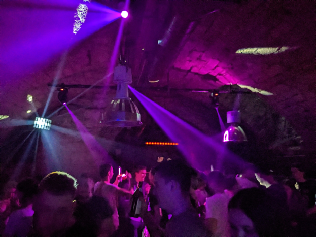 Gruppe von Menschen, die in einem Nachtclub tanzen, mit lila und blauen Deckenlichtern, einige halten Flaschen.