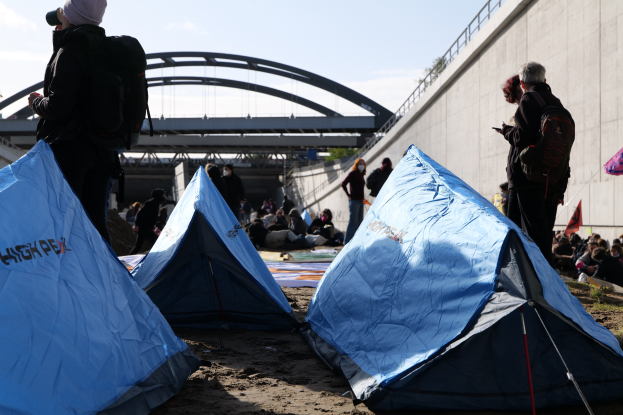 Eine Gruppe von Menschen sitzt auf einem sandigen Strand in der Nähe von Zelten, mit einer Wand und einer Brücke im Hintergrund, die an einer Klimawandeldemonstration teilnehmen.