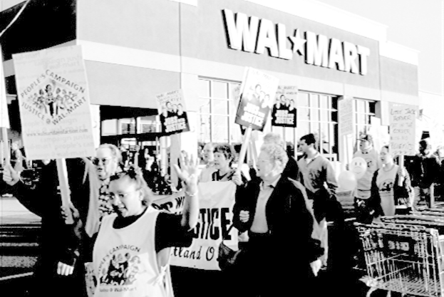 Ein Schwarzweißbild von protestierenden Menschen mit Plakaten und einem Einkaufszentrum im Hintergrund.