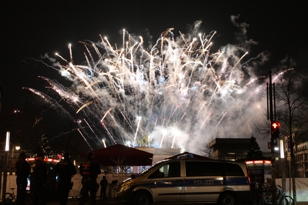 Ein Polizeiwagen steht vor einer Menschenmenge, mit beleuchteten Gebäuden, Bäumen, Masten und Verkehrszeichen im Hintergrund, unter einem Himmel voller farbiger Feuerwerke während einer Silvesterfeier in Berlin.