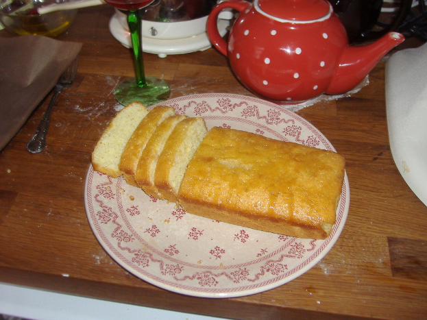 Ein weißer Teller mit Brot auf einer braunen Oberfläche, mit einem Glas, einem Kessel und einem weißen Objekt oben, einer Gabel und einem Tablett links, einer Schüssel in der oberen linken Ecke und einem weiteren weißen Objekt rechts.