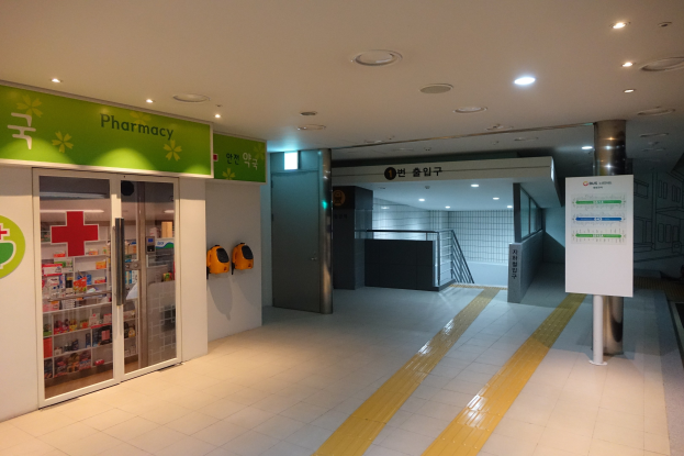 Apotheken-Eingang mit einem "Apotheke"-Schild auf der linken Seite, einer Tafel mit Text auf der rechten Seite, Hintergrundobjekte und beleuchteter Dachfirst.