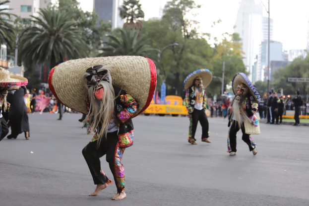 Eine Gruppe von Menschen in bunten Kostümen und Sombreros tanzt eine Straße mit Bäumen und Laternen in Mexiko-Stadt hinunter, mit Gebäuden und Strommasten im Hintergrund.