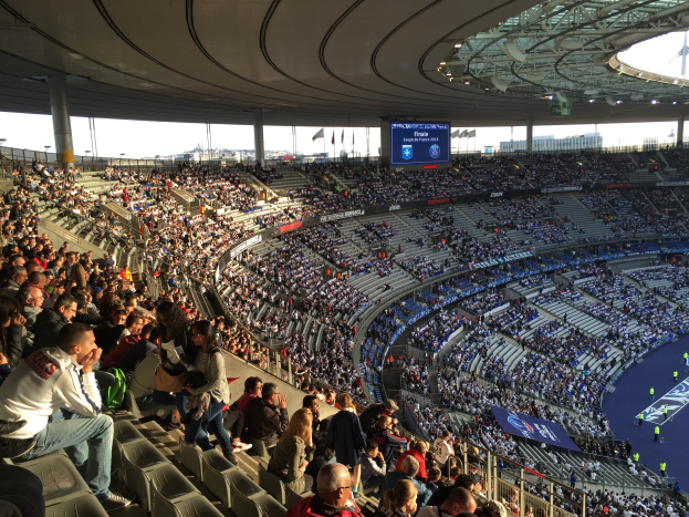 Große Zuschauermenge in einem Stadion bei einem Fußballspiel, mit einer Bühne rechts, Fahnen, Stangen, einem Bildschirm und dem Allianz Stadion in München, Deutschland im Hintergrund.