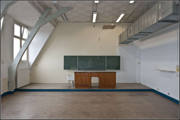 Ein leeres Klassenzimmer mit einer Tafel, einem Tisch, einem Waschbecken mit Wasserhahn, einem Hocker, einem Fenster und Deckenlampen.