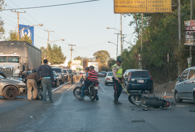 Gruppe von Menschen um ein verunglücktes Motorrad auf dem Seitenstreifen mit mehreren Fahrzeugen, darunter ein Lastwagen, im Hintergrund und Bäumen, Masten, Lichtern, Schildern und Himmel.