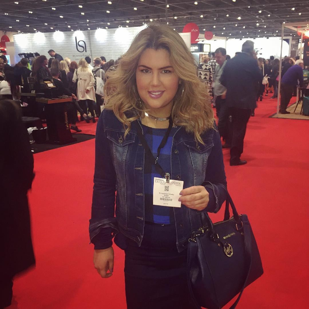 Frau auf einem roten Teppich bei einer Modenschau, die lächelt und eine Handtasche hält, mit Fotografen und Lichtern im Hintergrund.