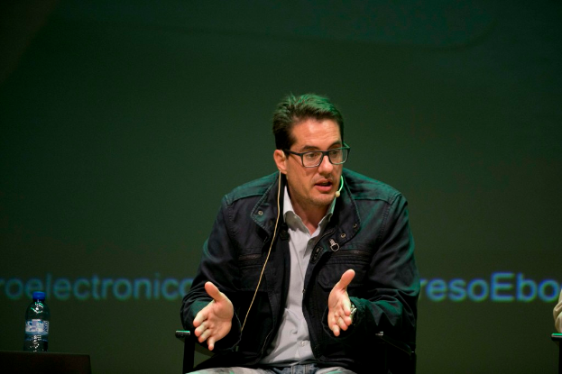 Eine Person sitzt in der Mitte auf einem Stuhl und hält eine Rede mit einem Mikrofon, während eine Flasche mit einer blauen Kappe links auf dem Tisch steht und ein Bildschirm im Hintergrund zu sehen ist.