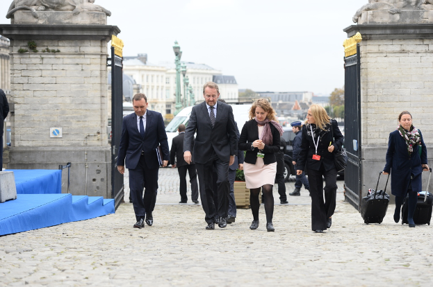 Eine Gruppe von Mitarbeitern geht in einen Bereich mit einem Tor, hinter dem sich viele Fahrzeuge und Gebäude befinden, während eine Frau auf der rechten Seite zwei Gepäckstücke trägt.