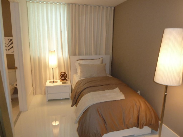 Ein Zimmer mit einem Bett, zwei Kissen, einem Nachttisch mit Gegenständen, einem Handtuch, einer Papierrolle, an der Wand angebrachten Gegenständen, zwei Lampen, einem Vorhang und einem sichtbaren Badezimmer.