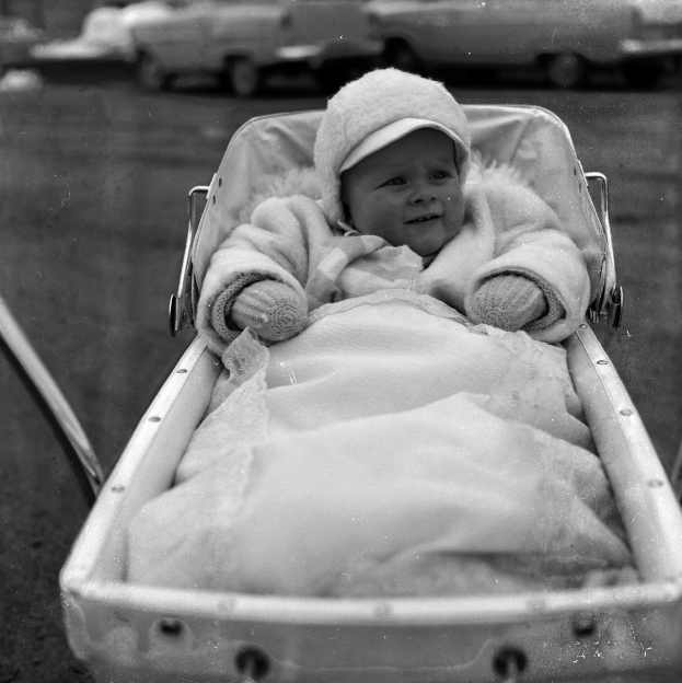 Ein Schwarz-Weiß-Foto eines lächelnden Babys in einem Kinderwagen, das eine weiße Kleidung und eine Kappe trägt, mit Fahrzeugen im Hintergrund.