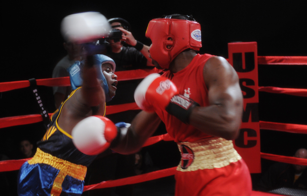 Zwei Boxer in einem Ring, die in einem Kampf gegeneinander antreten, mit einem Fotografen und Zuschauern im Hintergrund bei gedämpfter Beleuchtung.