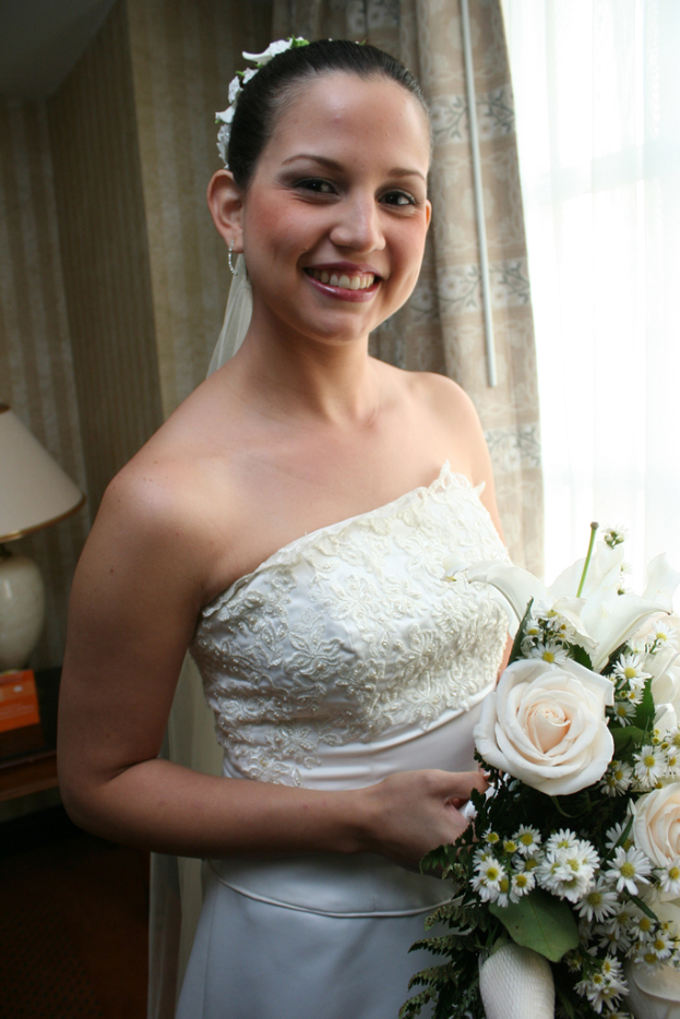 Eine strahlende Braut mit einem Bouquet in der Hand, vor einem Vorhang und einer Wand im Hintergrund und einer Lampe auf einem Tisch links