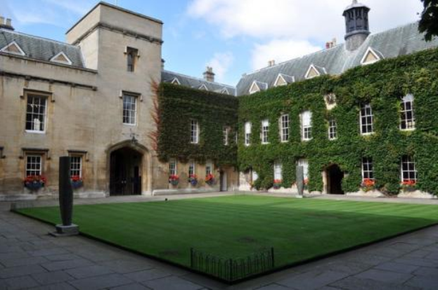 Die Universität Oxford, ein großes Gebäude mit Efeu an seinen Wänden, Fenstern und einer Tür, umgeben von grünem Gras und blühenden Pflanzen, unter einem bewölkten Himmel.