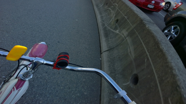 Ein Fahrradlenker ist unten auf dem Bild zu sehen, mit Fahrzeugen, die auf der Straße rechts fahren und einer Trennung in der Mitte.