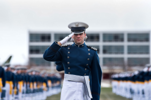 Ein Mann in militärischer Uniform salutiert auf einer Abschlussfeier und ist von einer Menge umgeben, mit einem Gebäude und Himmel im Hintergrund.