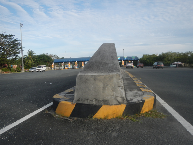 Ein Betonbollard an der Seite einer Straße mit vorbeifahrenden Autos, Bäumen, Polen, Schuppen und einem klaren blauen Himmel im Hintergrund.