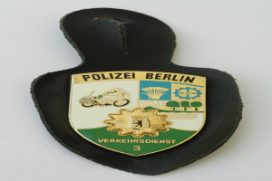 Ein Polizeistern mit der Aufschrift "Polizei Berlin" darauf, der ein blaues Schild mit einem weißen Stern in der Mitte zeigt, umgeben von einem weißen Rand, auf einer Oberfläche platziert.
