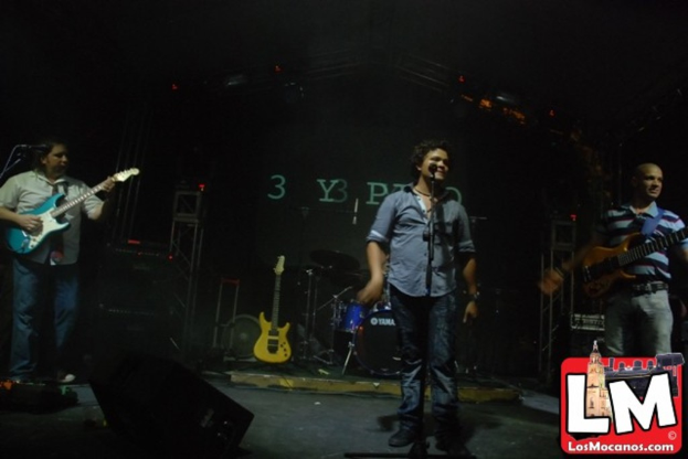 Drei Personen auf der Bühne, eine spielt Gitarre und eine singt in ein Mikrofon, mit einem Snaredrum-Arrangement und Beleuchtung im Hintergrund.