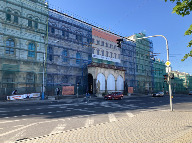 Großes Gebäude mit Gerüst während der Renovierung umgeben von Fahrzeugen, Passanten, Ampeln, Schildern, Straßeninfrastruktur, Bäumen und einem klaren blauen Himmel.