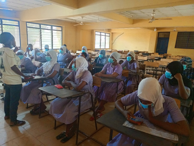 Eine Klasse in Nigeria mit Schülern, die Gesichtsmasken tragen und auf Bänken sitzen oder stehen, mit Büchern und Gegenständen auf Tischen, Fenstern, einer Tür, einer Wand, einer Decke mit Lampen und Ventilatoren im Hintergrund.