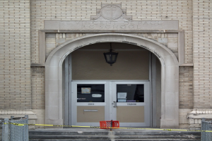 Gelbes Absperrband umgibt den Eingang eines Backsteingebäudes mit einer mit Papieren bedeckten Tür, Treppe mit Maschendrahtzäunen auf beiden Seiten und einer Deckenlampe; die Wand des Gebäudes zeigt wahrscheinlich den Namen der Schule.
