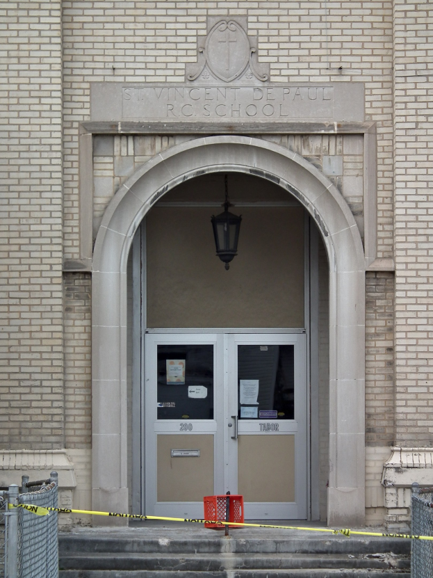 Gelbes Absperrband umgibt den Eingang eines Backsteingebäudes mit einer mit Papieren bedeckten Tür, Treppe mit Maschendrahtzäunen auf beiden Seiten und einer Deckenlampe; die Wand des Gebäudes zeigt wahrscheinlich den Namen der Schule.