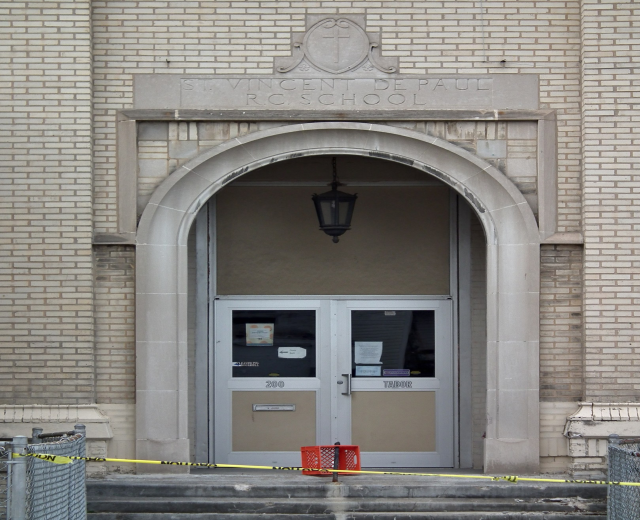 Gelbes Absperrband umgibt den Eingang eines Backsteingebäudes mit einer mit Papieren bedeckten Tür, Treppe mit Maschendrahtzäunen auf beiden Seiten und einer Deckenlampe; die Wand des Gebäudes zeigt wahrscheinlich den Namen der Schule.