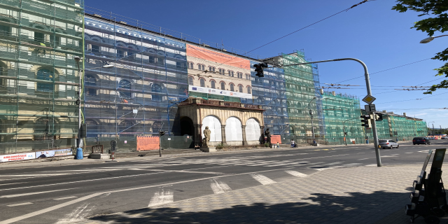 Großer mehrgeschossiger Bau unter construction in Berlin mit zahlreichen Fenstern, umgeben von Straßeninfrastruktur, Fahrzeugen, Fußgängern und Bäumen unter einem bewölkten Himmel.