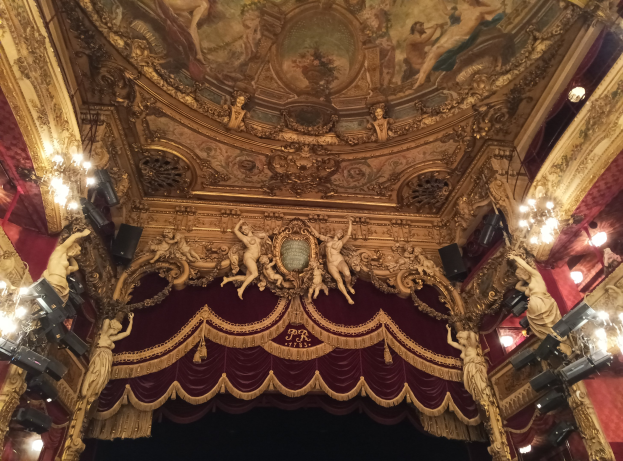 Theaterinnenraum mit roter Gardine, Kronleuchtern, Deckenplastiken, Lichtern und Gemälde, mit einer Bühne mit einigen Menschen unten.