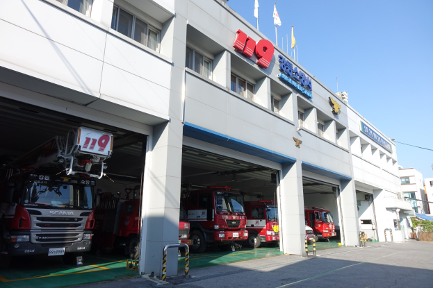 Feuerwache mit mehreren Feuerwehrautos vor der Tür, mit Fenstern, Säulen und Namensschildern, Flaggen im Vordergrund, anderen Gebäuden im Hintergrund und einem klaren blauen Himmel.