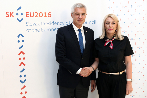 Zwei Personen, die nebeneinander stehen und lächeln, der Mann trägt einen schwarzen Blazer mit einem weißen Hemd und einer blauen Krawatte, die Frau trägt eine Brille, mit "sk eu 2016" auf einer Tafel dahinter.