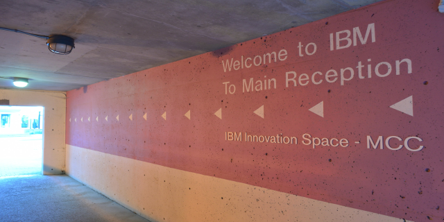 Eine Wand Mit Den Wörtern "Willkommen Bei IBM" Beleuchtet Von Deckenlampen, Mit Einer Tür Im Hintergrund, Die Zum IBM Innovation Space MCC Führt.