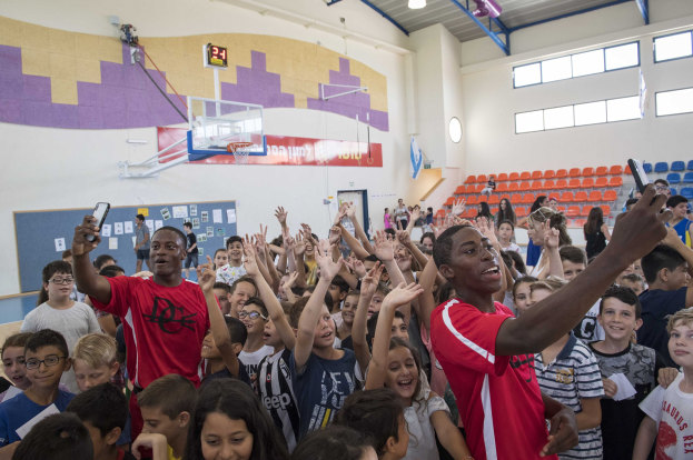 Kinder vor einem Basketballfeld mit Handys stehend, mit einer Pinnwand, Uhr, Torpfosten, Basketballnetz, Deckenleuchten, Stühlen und Fenstern im Hintergrund auf einem Basketballcamp.