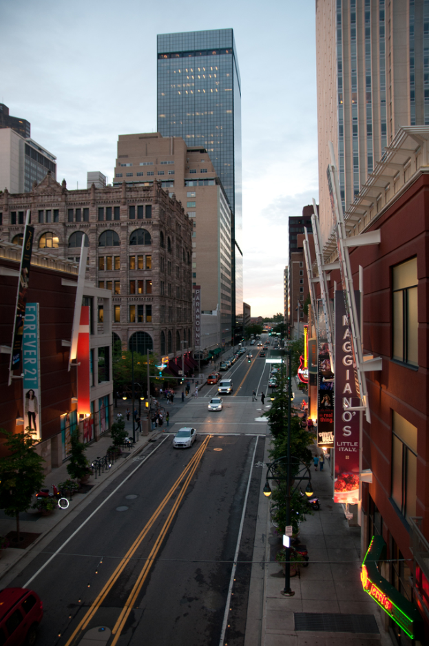 Stadtansicht mit einer Straße voller Autos unten, flankiert von Gebäuden und Wolkenkratzern auf beiden Seiten, unter einem Himmel oben.