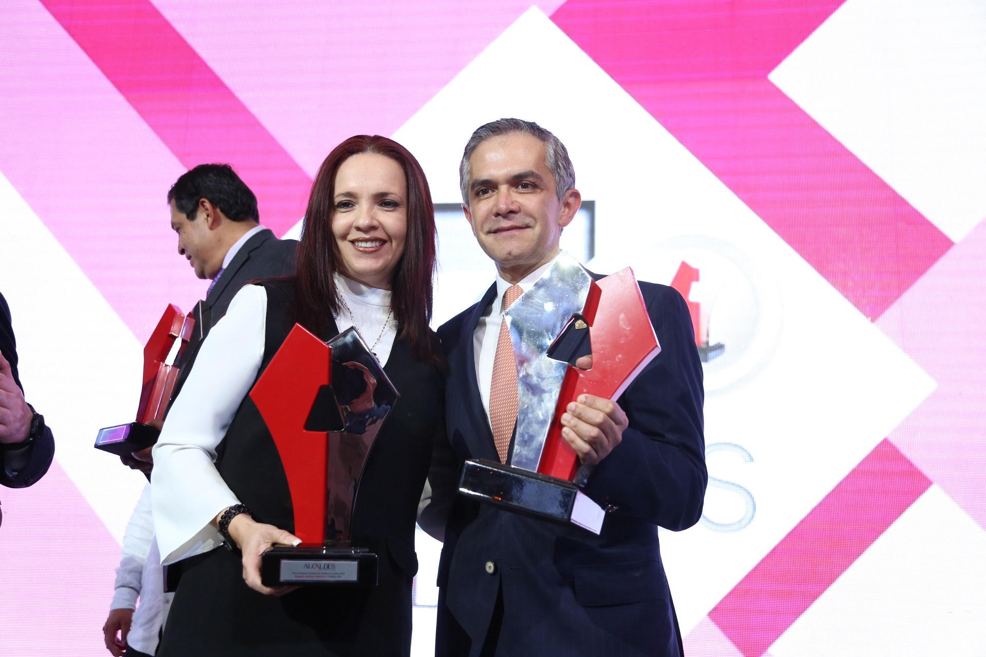 Zwei Personen auf einer Bühne, die lächeln und Pokale halten, mit einem sichtbaren Bildschirm dahinter.