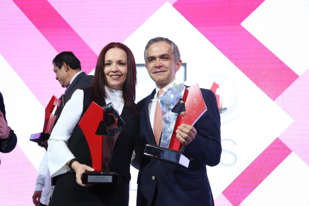 Zwei Personen auf einer Bühne, die lächeln und Pokale halten, mit einem sichtbaren Bildschirm dahinter.