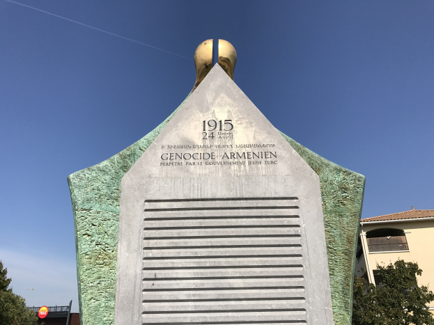 Ein hoher Denkmal mit einer Uhr darauf, mit Text beschriftet, steht in einem Park umgeben von Bäumen und Gebäuden unter einem klaren Himmel, zum Gedenken an den 24. Jahrestag des Völkermords in Armenien.