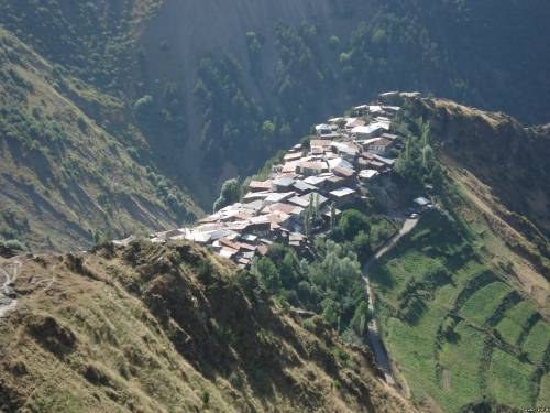 Kleines Dorf mit Häusern, die zwischen saftigen grünen Hügeln und Bäumen in einer Berglandschaft liegen.