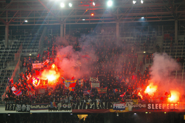 Eine große Menschenmenge in einem Stadion hält Fahnen und Banner, mit Leuchtraketen und Rauch, der aus ihnen aufsteigt, während Metallrahmen und Deckenleuchten darüber sichtbar sind.