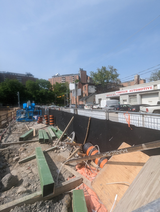 Baustelle mit verschiedenen Materialien wie Holzplanken, Rohren und Steinen, umgeben von Fahrzeugen, Gebäuden, Bäumen und einem klaren blauen Himmel.