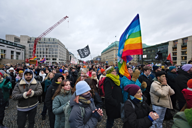 Eine große Gruppe von Menschen bei einer LGBTQ+-Rechtsdemo in Berlin, die Fahnen und Plakate schwenkt, mit Gebäuden und einem Kran im Hintergrund.