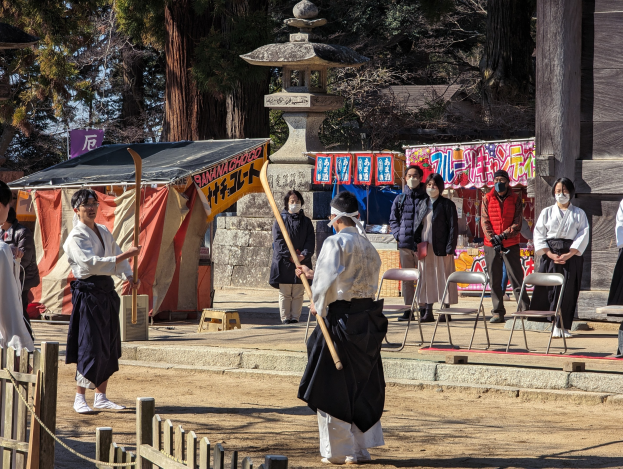 Eine Gruppe von Menschen in traditioneller Kleidung versammelt sich im Freien in Kyoto, einige tragen Masken und halten Holzstöcke, mit Stühlen, Bannern, einem Zelt und einem Holzzaun gegen einen klaren blauen Himmel.