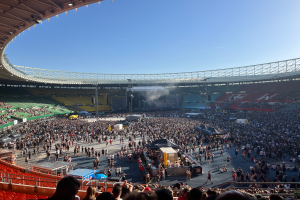 Eine große Menschenmenge versammelte sich im Wembley-Stadion für ein Konzert, mit einer Bühne in der Mitte und einem Dach mit Lichtern an der Spitze des Bildes.