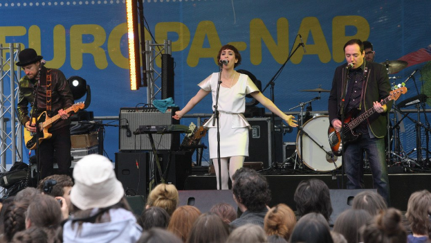Vier Personen, die auf einer Bühne Musik spielen, mit einer Frau in einem weißen Kleid, die singt, und zwei Männern, die Gitarren spielen, während eine Menge davor steht und eine Wand mit Lautsprechern im Hintergrund zu sehen ist.